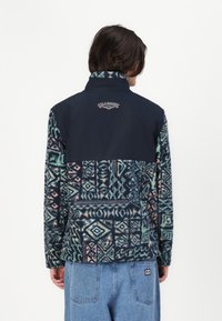 Fleece striukė su geometriniais raštais turkio, rožinės ir tamsiai mėlynos spalvų. Yra tvirta tamsiai mėlyna viršutinė dalis ir aukšto apykaklės dizainas.
