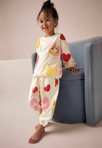Completo pigiama per bambini in crema con motivi di cuori colorati. La parte superiore presenta un'illustrazione di un personaggio dei cartoni animati; i pantaloni hanno polsini elastici.
