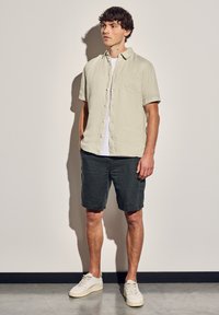 Camisa de lino gris clara de manga corta con un bolsillo en el pecho, combinada con pantalones cortos gris oscuro y zapatillas blancas, sobre un fondo liso.