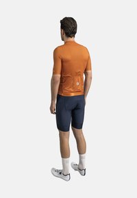 Oranje fietsshirt met korte mouwen van gestructureerde stof, achterzakken; gecombineerd met aansluitende marineblauwe shorts en witte fietsschoenen.