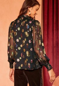 Blusa nera con maniche trasparenti decorate da pois multicolori e lucenti. Presenta maniche a sbuffo e una chiusura con bottoni al collo.