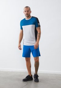 Athletisk T-skjorte i blå, hvit og svart med korte ermer; kombinert med blå shorts. Har en logo og stripete detaljer på ermet.
