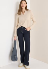 Beżowa bluzka z długim rękawem i dekoltem w serek, zestawiona z granatowymi dzwonami z kontrastującym przeszyciem. Szara torba i białe trampki dopełniają stylizację.