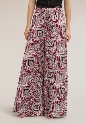 Pantalon à jambes larges avec motifs floraux et géométriques abstraits rouges, blancs et noirs, porté avec des sandales ouvertes et un haut noir.
