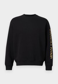 Melnā kapuce, izgatavota no kokvilnas maisījuma, ar apaļu kakla izgriezumu un ribotiem piedurknēm. Zelta "ARMANI EXCHANGE" logo uzdrukāts gar kreiso piedurkni.