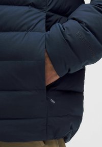 Tengerészsötét pufidzseki steppelt mintával, cipzáras zsebbel és logóval a hónalján. Az anyag simának és puhanak tűnik.