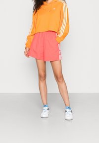 Person, der einen orangefarbenen Adidas-Hoodie und korallenfarbene Adidas-Shorts mit weißen Streifen trägt, steht mit einer Hand in der Tasche und weißen Turnschuhen.