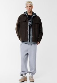 Giacca oversize marrone in lana sopra una felpa grigia con cappuccio, camicia a quadri, pantaloni della tuta grigi e sneakers beige. Presenta due tasche anteriori.