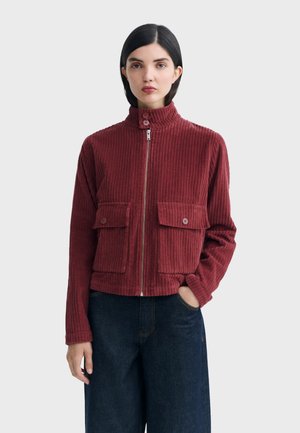 Femme portant une veste zippée côtelée bordeaux avec col montant et grandes poches à boutons, associée à un jean large bleu foncé.