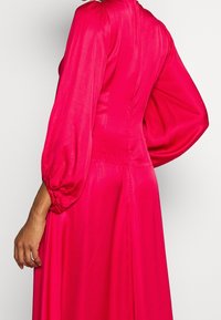 Kvinna i en klar rosa satin klänning med biskopsärm och dragkedja i ryggen, sedd från sidan och bakifrån.