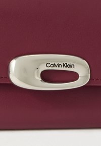 Bordeaux leren oppervlak met een zilver metalen sluiting met een rechthoekige uitsparing en "Calvin Klein" erin gegraveerd.