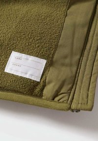 Pochette vert olive avec un panneau en polaire texturé et un tissu lisse. Comprend une étiquette blanche pour indiquer le nom et les coordonnées téléphoniques.