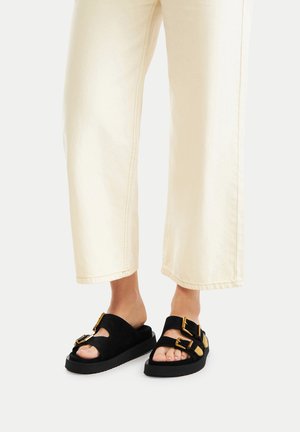 Schwarze Wildleder-Sandalen mit zwei verstellbaren Riemen, goldfarbenen Metallakzenten und einer dicken, strukturierten schwarzen Sohle, kombiniert mit beigen Hosen mit weitem Bein.