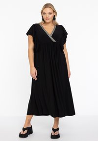 Schwarzes Maxikleid mit V-Ausschnitt und kurzen, schwingenden Ärmeln, das mit einem dekorativen Band am Ausschnitt versehen ist. Das Model kombiniert es mit klobigen schwarzen Plateausandalen.