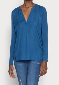 Blå långärmad topp med V-ringning, som har en avslappnad passform och mjuk textur. Anmärkningsvärda detaljer inkluderar en knappdekoration vid halslinningen.