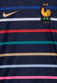 Jersey azul marino con rayas horizontales en rojo, azul, verde azulado, blanco, rosa claro y beige. Presenta un logotipo dorado de Nike y un emblema de gallo bordado.