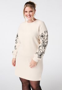 Crème jurk met zwarte bloemenborduursels op de mouwen, lange losse pasvorm, ronde halslijn en zijsplitten aan de onderrand. Glad stoftextuur.