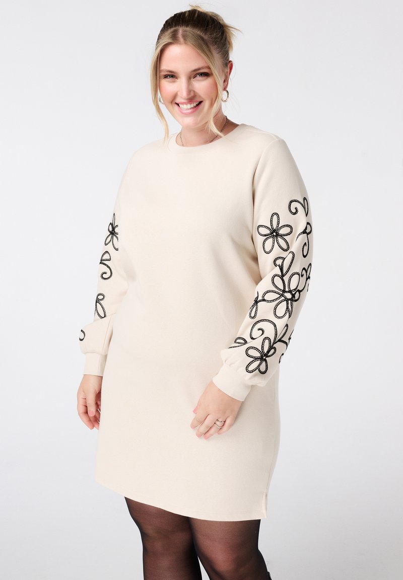 Crème jurk met zwarte bloemenborduursels op de mouwen, lange losse pasvorm, ronde halslijn en zijsplitten aan de onderrand. Glad stoftextuur.