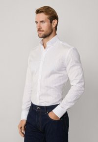 Biela košeľa na gombíky s ružovými pruhmi, s dlhými rukávmi a špicatým golierom, v kombinácii s tmavomodrými džínsami. Hladká tkanina.