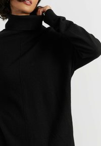 Zwarte coltrui van zacht materiaal, met een relaxte pasvorm en geribbelde manchetten en halslijn, die een naadloos ontwerp benadrukt.