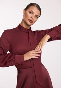 Bordowa sukienka z wysokim dekoltem, detalem w postaci kokardy i bufkami; wykonana z gładkiego materiału z guziczkami na mankietach.