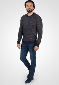 Marineblauer gestrickter Pullover mit diagonalem Muster, gerippten Bündchen und Saum, kombiniert mit schmal geschnittenen blauen Jeans und marinefarbenen Sneakern. Das Model steht vor einem hellen Hintergrund.