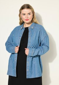 Camicetta in denim blu con bottoni e dettagli in perle, maniche lunghe e colletto, indossata sopra un vestito nero.