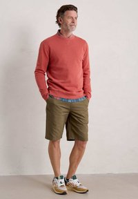 Felpa di un leggero corallo, pantaloni corti verdi e sneakers multicolori. L'outfit presenta un look a strati con una camicia a fantasia sotto la felpa.