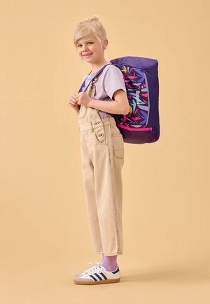 Lila Rucksack mit bunten Blattmustern, getragen über einem helllila T-Shirt und beigen Latzhosen, kombiniert mit Sneakers und lila Socken.