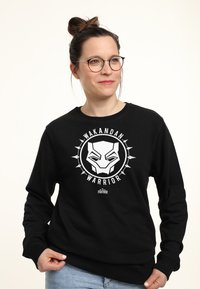 Czarna bluza z widocznym białym graficznym wizerunkiem maski pantery z napisem "Wakanda Wojownik", otoczona okrągłym wzorem i kolcami.