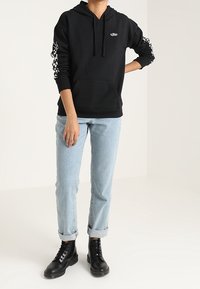 Sudadera negra con un bolsillo frontal, que presenta un logo blanco y acentos con patrones en las mangas. Combinada con unos jeans azules claros remangados y botas negras.