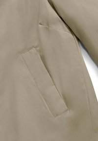Giacca leggera beige con una texture liscia. Presenta un colletto ripiegato e una tasca laterale a sinistra. Design semplice, senza motivi visibili.