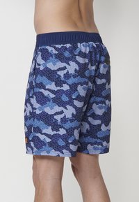 Modré camo plavky s geometrickým vzorom. Elastický pás s kontrastným námorníckym dokončením a oranžovým logom pri leme.