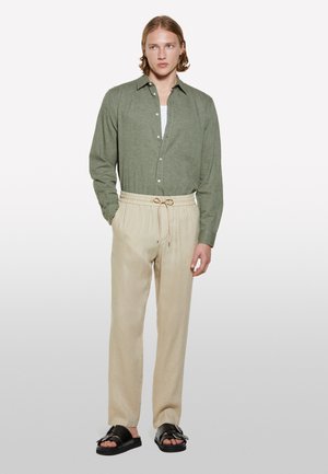 Chemise verte à manches longues et boutonnage, associée à un pantalon beige à cordon de serrage et des sandales noires. Coupe simple et décontractée avec un minimum de détails.