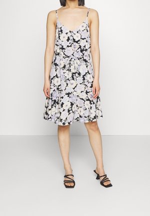 Kobieta w czarnej, kwiecistej sukience bez rękawów z wiązaniem w talii i czarnych sandałach na obcasie, stojąca na jednolitym jasnym tle.