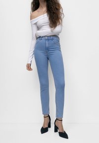 Jean taille haute bleu clair avec une coupe slim, associés à un haut à manches longues blanc dénudé des épaules et des chaussures à talons noires.