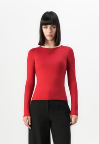 Vrouw met steil zwart haar, gekleed in een strakke rode lange mouwtop en losse zwarte broek, staand tegen een effen witte achtergrond.