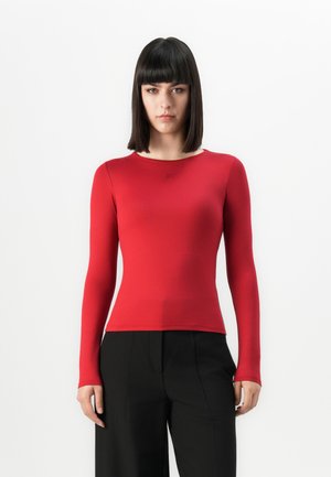 Woman with straight black hair wearing a fitted long-sleeve red top and loose black pants, standing against a plain white background.