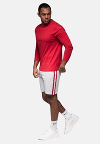 Camisa roja de manga larga combinada con pantalones cortos grises que tienen rayas rojas a los lados. Completan el look unas zapatillas blancas y calcetines hasta el tobillo.