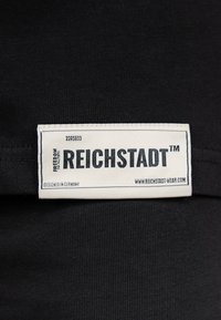 Zwart stoffen label met witte tekst. Er staat "REICHSTADT™" op, samen met "FREEDOM", "DESIGNED IN GERMANY" en een website. Eenvoudig, rechthoekig ontwerp.