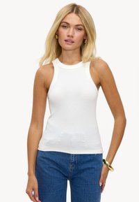 Débardeur blanc à côtes avec col montant et sans manches, associé à un jean en denim bleu. Texture lisse, forme ajustée.