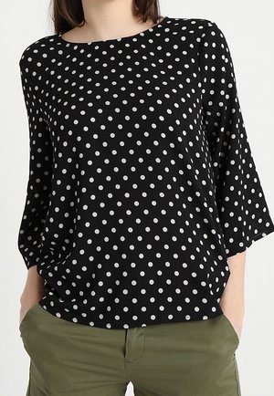 Chemise noire à manches longues avec des pois blancs, portée avec un pantalon vert olive, mains partiellement dans les poches, tête partiellement hors du cadre.