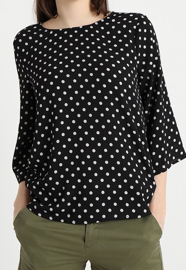 Svart långärmad tröja med vita prickar, iklädd olivgröna byxor, händerna delvis i fickorna, huvudet delvis utanför ramen.