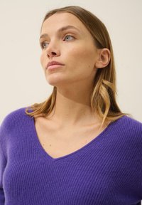 Fioletowy wełniany sweter z dekoltem w szpic, z teksturowanym ściągaczem. Materiał wydaje się miękki i lekko grubszy w strukturze.