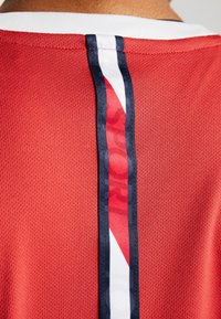 Červený sportovní dres zezadu s vertikálním bílým a navy pruhy, ve kterém je blízko krku částečně viditelný červený text.