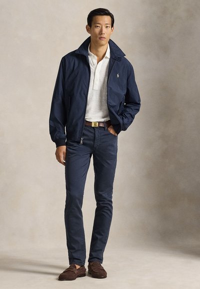 Άνδρας που στέκεται φορώντας μπλε σκούρο σακάκι, λευκό πόλο πουκάμισο, μπλε σκούρο παντελόνι, καφέ ζώνη και καφέ σουέντ μοκασίνια, με φόντο απλό.