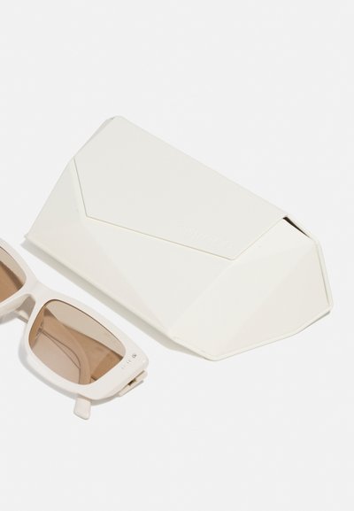 Gafas de sol blancas con un marco geométrico y lentes marrones al lado de un estuche angular blanco a juego, con un sutil branding y una superficie texturizada.