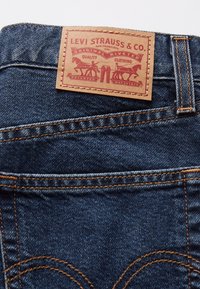 Jean en denim bleu foncé avec des coutures orange et une étiquette en cuir marron portant le logo et le texte de Levi Strauss & Co.