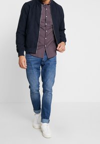 Giacca bomber blu navy con zip, camicia bordeaux a motivo sotto, jeans azzurri e sneakers bianche. Design casual e aderente.