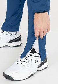 Sportbyxor i marinblått med dragkedjor vid vristerna, kombinerat med vita sneakers med svarta och grå detaljer samt nätstruktur.